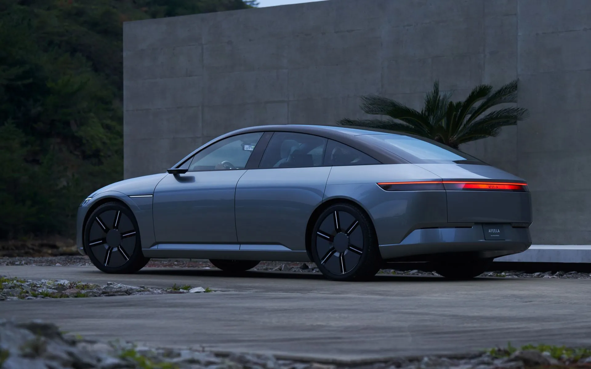 Elektrische sedan in schemerlicht voor een betonnen muur, zijaanzicht met doorlopende rode achterlichtbalk