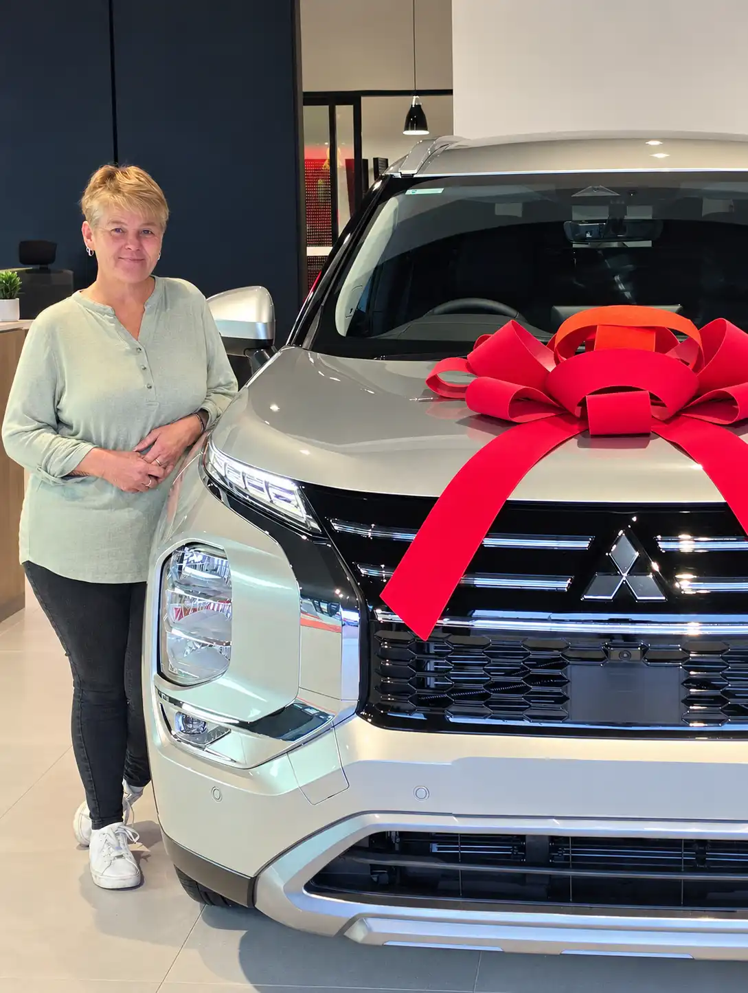 Tevreden klant leunt tegen haar nieuwe zilverkleurige Mitsubishi Outlander met een grote rode strik op de motorkap